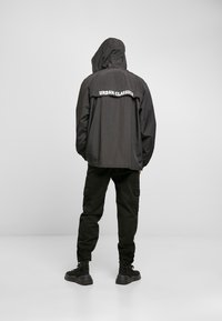 Black lightweight jacket with a hood, featuring "URBAN CLASSICS" logo across the upper back; paired with black cargo pants and boots.