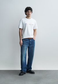 Mladý muž s kučeravými vlasmi, nosiaci okuliare, bielu tričko s potlačou, modré džínsy, hnedý opasok a čierne topánky, stojaci pred jednofarebnou sivou stenou.