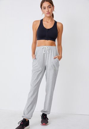 Kobieta stojąca z rękami w kieszeniach, ubrana w czarny sportowy biustonosz na zamek, jasnoszare spodnie dresowe ze sznurkiem oraz czarne buty sportowe.