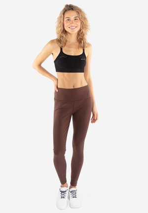 Młoda kobieta uśmiecha się, stojąc z jedną ręką na biodrze, ubrana w czarny sportowy biustonosz, brązowe legginsy i białe buty sportowe na białym tle.