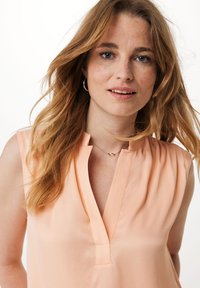 Bluzka bez rękawów w kolorze brzoskwiniowym z dekoltem w kształcie litery V, wykonana z miękkiego materiału, z delikatnymi plisami, uzupełniona subtelnym złotym naszyjnikiem.