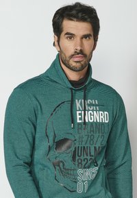Tirkizna hoodie s teksturiranom površinom, s grafikom lubanje i bijelim tekstualnim detaljima. Uže na vratu upotpunjuje dizajn.