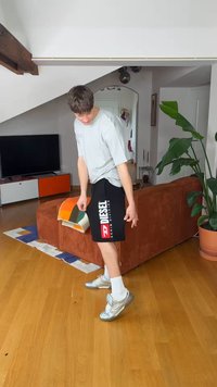 Jeune homme portant un short Diesel et des baskets argentées, debout sur un sol en bois dans un salon avec un canapé marron et une plante verte.