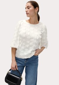 Witte textuurblouse met bloemenapplicaties en 3/4 mouwen, gedragen met blauwe jeans en een kleine zwarte handtas.