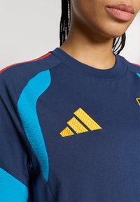 Μπλε ναυτικό αθλητικό πουκάμισο με τιρκουάζ και κόκκινες λεπτομέρειες, με κίτρινο κεντημένο λογότυπο. Το υλικό φαίνεται υφασμάτινο με κοντά μανίκια.