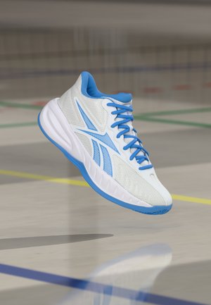 Scarpa sportiva bianca e blu con lacci sospesa sopra un parquet lucido da palestra con linee colorate del campo.