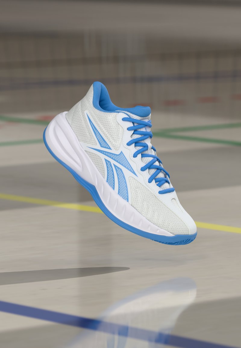 Λευκό και μπλε αθλητικό παπούτσι με κορδόνια να αιωρείται πάνω από ένα γυαλισμένο εσωτερικό γήπεδο με έγχρωμες γραμμές γηπέδου.
