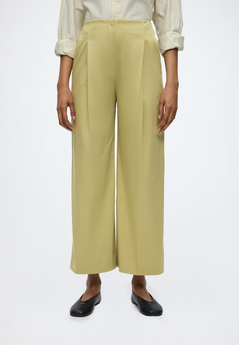 Femme portant un pantalon beige taille haute à jambes larges, une chemise à manches longues rayée avec les manches retroussées, et des ballerines noires, debout devant un fond blanc.