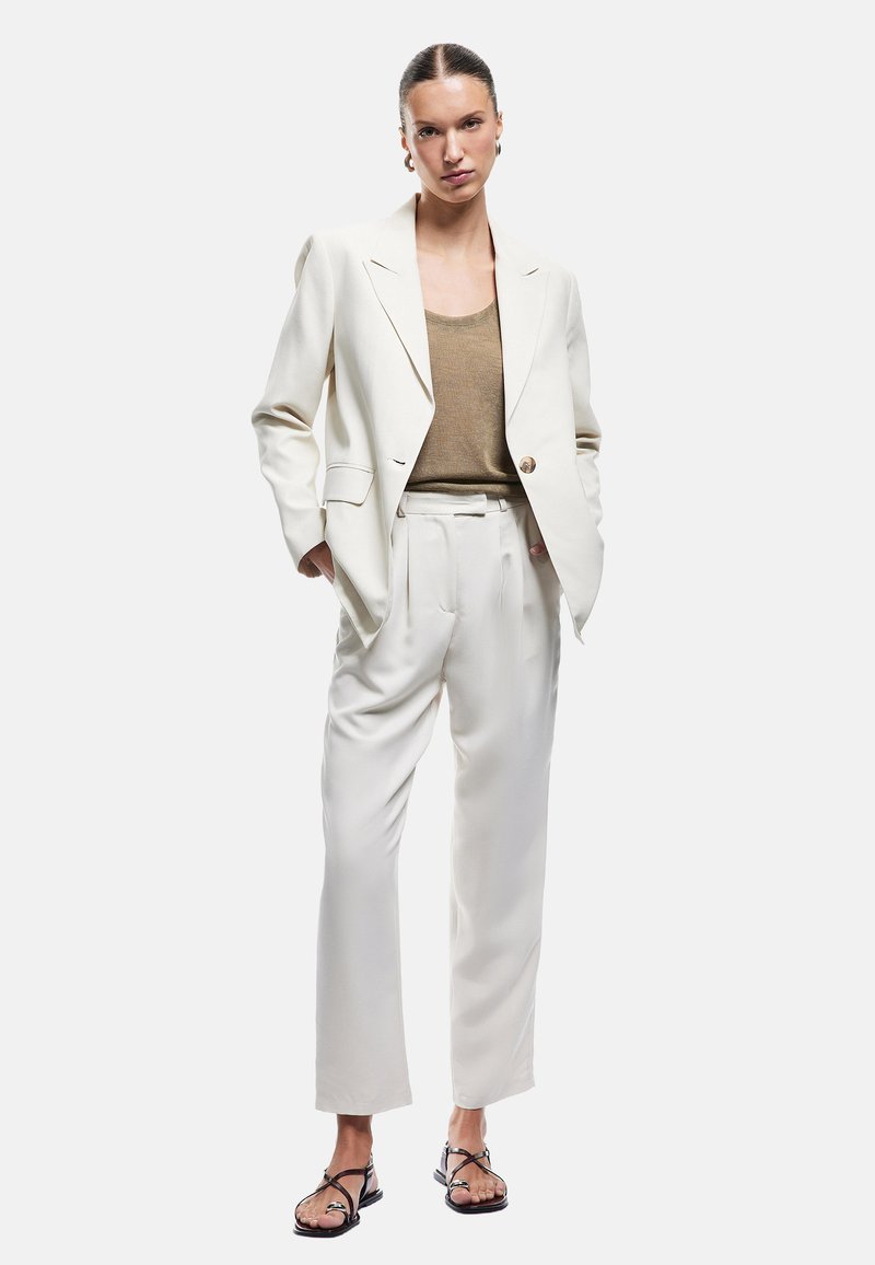 Blazer i cremefarvet stof med stiklapeller, parret med en lysebrun top og hvide bukser. Modellen har sorte sandaler på med tynde stropper.