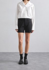 Chemise blanche à col avec une poche poitrine, associée à un short noir taille haute avec des boutons sur le côté. Chaussettes noires et mocassins brillants complètent le look.