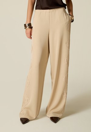 Pantalon beige à jambes larges avec des détails en dentelle, porté avec des chaussures noires pointues, un haut noir et des bracelets dorés à un poignet.