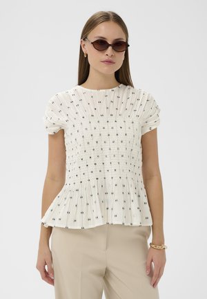 Biała bluzka z marszczonym stanem, krótkimi bufkami i czarnym wzorem w kropki. Rozkloszowany dół typu peplum doskonale komponuje się z beżowymi spodniami.