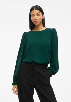 Donna con capelli lunghi e scuri, che indossa una blusa lunga a maniche lunghe di un verde scuro texturizzato e pantaloni neri a vita alta, in piedi con una mano in tasca.
