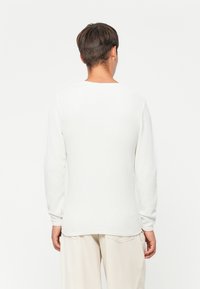 Witte longsleeve shirt met een gestructureerd, geribbeld patroon en ronde halslijn, weergegeven vanuit de achterkant, gecombineerd met lichte broek.
