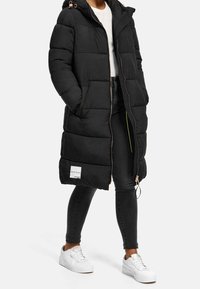 Svart puffjacka med huva, dragkedja fram och elastiska detaljer. Har en knälång design, sidofickor och en varumärkeslapp.