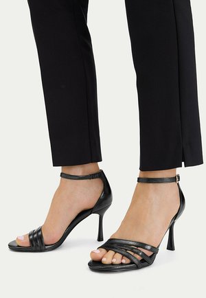 Piedi con sandali neri con tacco alto e cinturini alla caviglia, abbinati a pantaloni neri slim fit con spacchi frontali.