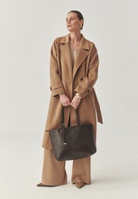 Brun yllekappa, beige topp, bruna vida byxor och spetsiga skor. Svart handväska med krokodilmönster och långa handtag. Guldaccessoarer.