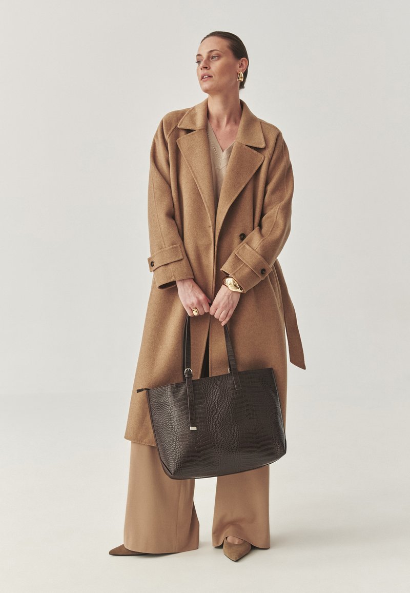 Brun yllekappa, beige topp, bruna vida byxor och spetsiga skor. Svart handväska med krokodilmönster och långa handtag. Guldaccessoarer.