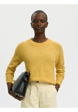 Osoba ubrana w żółty sweter i kremowe spodnie, trzymająca czarną skórzaną torebkę, stojąca na tle jednolitego jasnego tła.