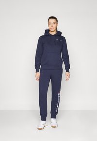 Ensemble de sweat à capuche et de pantalons de survêtement bleu marine en tissu doux. Le sweat à capuche possède une poche devant ; les pantalons présentent un graphique de logo sur le côté.