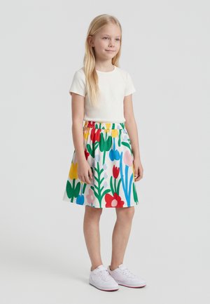 Fiatal lány áll, fehér bordázott rövid ujjú felsőt és színes virágos szoknyát visel, fehér tornacipővel, egyszerű háttér előtt.