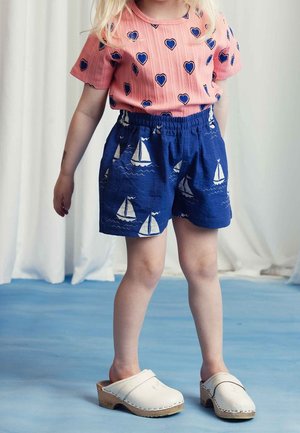 Enfant portant une chemise rose avec des cœurs bleus, un short bleu avec des imprimés de voiliers blancs, et des sabots en bois blancs se tenant sur un sol bleu.