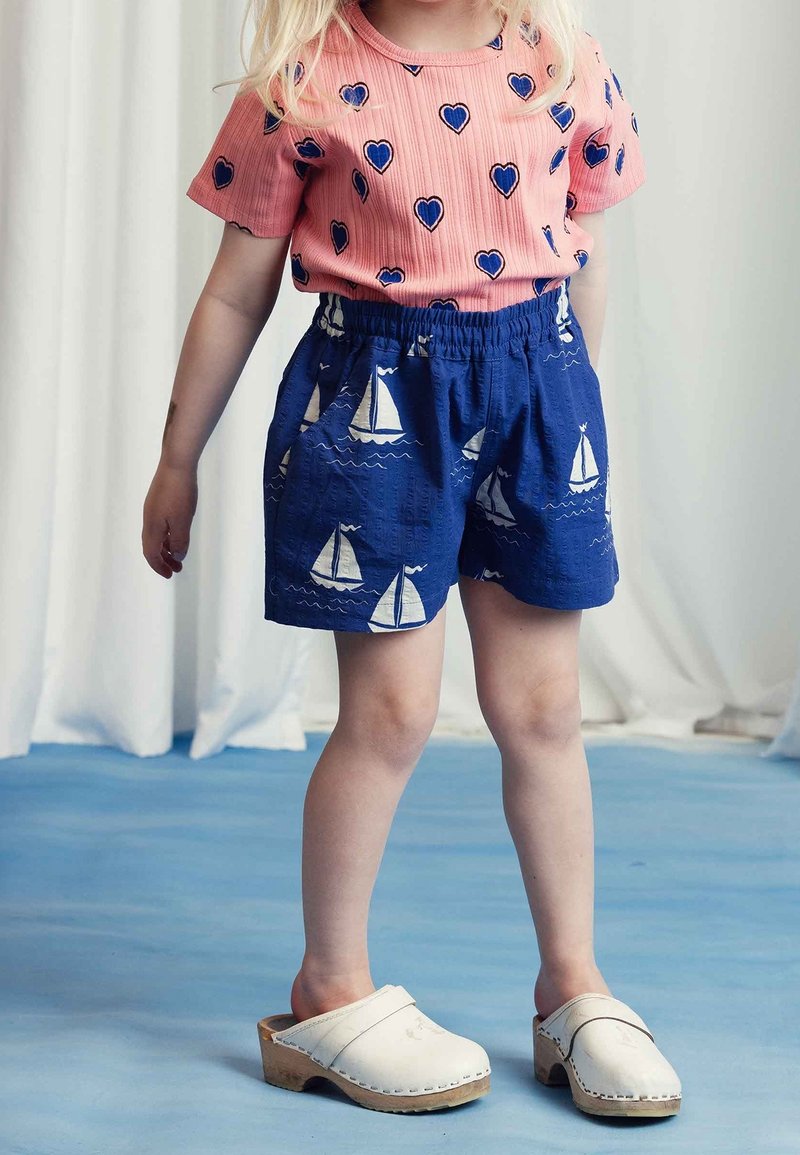 Enfant portant une chemise rose avec des cœurs bleus, un short bleu avec des imprimés de voiliers blancs, et des sabots en bois blancs se tenant sur un sol bleu.