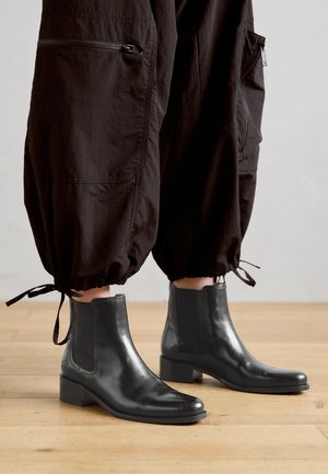 Sorte læder ankelstøvler med blokhæl og elastiske sidepaneler, parret med brune tætsiddende bukser med snore i bunden.