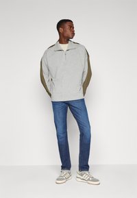 Light gray pullover with olive-green sleeves, half zipper, paired with blue jeans and gray sneakers with white stripes. Plain background.