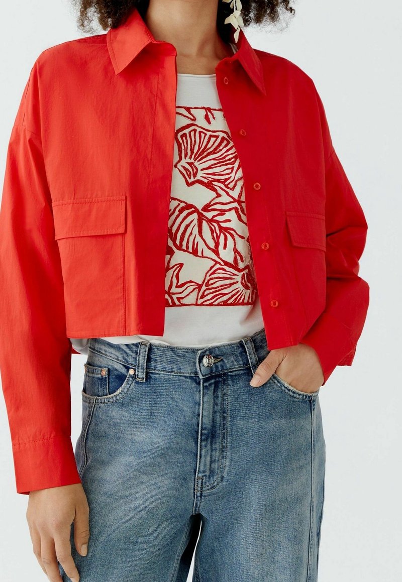 Personne portant une veste courte boutonnée rouge vif sur une chemise blanche avec un imprimé floral abstrait rouge et un jean en denim bleu avec la main dans la poche.