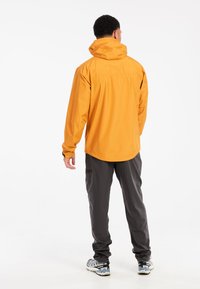 Man die met zijn rug naar ons toe staat, gekleed in een feloranje capuchonjack, donkergrijze broek en grijs-witte wandelschoenen.