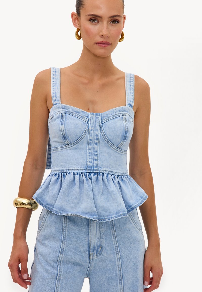 Femme portant un top peplum en denim bleu clair avec de larges bretelles et un pantalon en denim assorti, accessoirisée avec de grandes boucles d'oreilles en anneau dorées et un bracelet épais.