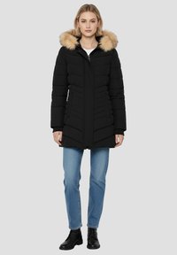 Veste matelassée noire avec une capuche doublée en fausse fourrure, dotée d'une fermeture éclair à l'avant, de coutures en biais et d'une coupe jusqu'aux genoux. Portée sur un jean bleu clair.