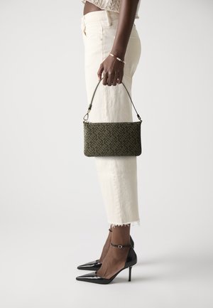 Borsa a spalla verde con motivo, dal design slim e con dettagli in oro, tenuta da una persona che indossa pantaloni bianchi e scarpe con tacchi neri.