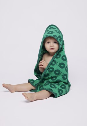 Malé dieťa sedí na podlahe s natiahnutými nohami, obalené v zelenom uteráku s kapucňou a tmavými kruhovými vzormi, pozerá sa dopredu so zloženými rukami.