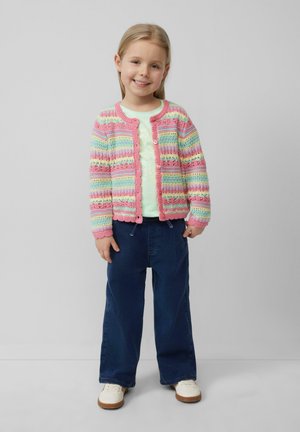 Kleurige, gebreide cardigan met roze, blauwe en gele strepen, met franjes aan de randen, gecombineerd met een lichtgroene top en donkerblauwe broek.