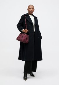 Abrigo largo negro de lana con botones de doble botonadura, combinado con un suéter blanco de canalé, pantalones negros y un bolso burdeos. Tacones negros.