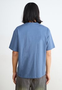 Tricou din bumbac albastru cu guler rotund, mâneci scurte și o croială lejeră. Textură netedă cu o culoare uniformă, fără modele sau detalii vizibile.