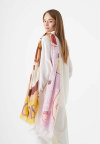 Kleurrijke, geprinte sjaal met roze, gele en bruine tinten, gemaakt van lichtgewicht stof, gedrapeerd over een witte blouse.