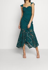 Teal spetsklänning med en figursydd livstycke, utfläkt nederkant med utskärningar och delikata axelrufsar. Sandal skor bärs med klänningen.