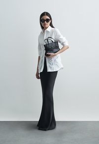 Λευκό πουκάμισο με κουμπιά και λεπτομέρειες με πιέτες, μαύρη στενή φούστα και μικρό μαύρο τσαντάκι. Το μοντέλο φοράει σκούρα γυαλιά ηλίου. Μινιμαλιστικό φόντο.