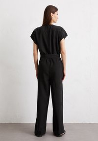 Zwarte jumpsuit met een getailleerde taille, korte mouwen en wijde benen. Heeft een gladde textuur, een rits aan de achterkant en minimale stikseldetails.