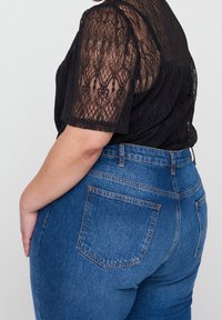 Top en dentelle noire à manches courtes, associé à un jean en denim bleu taille haute avec un design classique à cinq poches et des accents de couture jaune.