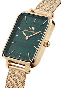 Fyrkantig klocka med en grönrandig urtavla och gyllene visare, med ett armband i guldtonat nät och varumärket "Daniel Wellington".