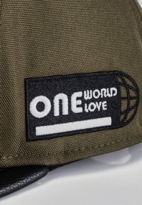 Gorra de tela verde olivo con un parche rectangular negro que lleva texto bordado en blanco: "UN MUNDO AMOR" y un gráfico de un globo. Visera negra.