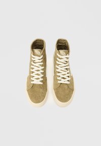 Υψηλές αθλητικές αθλητικές παπούτσια σε ελιά πράσινη σουέτ με άσπρα κορδόνια, τρυπητό δαχτυλο και κρέμας χρώματος καουτσούκ σόλα. Περιλαμβάνει λογότυπο στη γλώσσα.