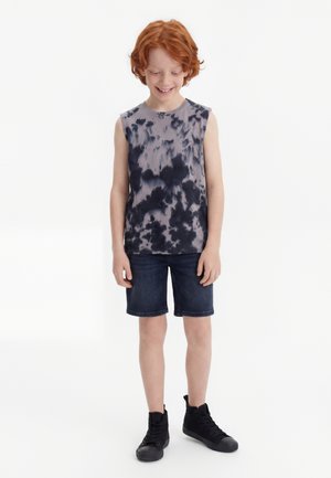 Garçon roux portant un débardeur tie-dye noir et gris, un short en jean foncé et des baskets montantes noires, souriant et regardant vers le bas.