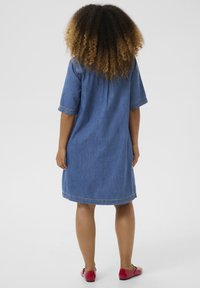Denim jurk in het blauw, middellang met korte mouwen, met een rechte snit en stiksels. Gstyled met rode platte schoenen.