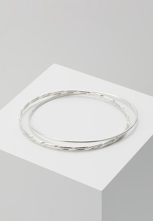 Zwei dünne silberne Armbänder, eines glatt und eines gedreht, flach liegend auf einem weißen quadratischen Podest vor grauem Hintergrund.