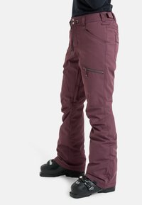 Bordeaux skibroek met een getailleerde pasvorm, ritszakken aan de zijkant, riemlussen en verstevigde zomen. Gecombineerd met zwarte skischchoenen.